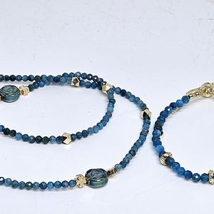 May include: A necklace and bracelet set with blue beaded strands. The necklace has gold-coloured clasps and accents, with a round clasp. The bracelet has a sliding clasp for adjustment.