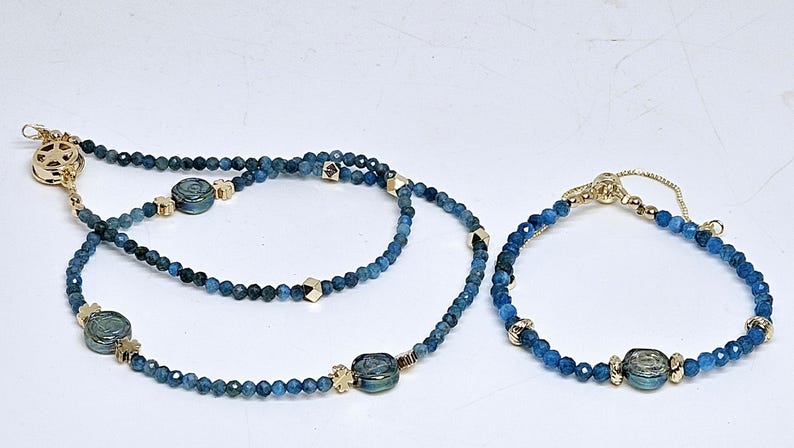 May include: A necklace and bracelet set with blue beaded strands. The necklace has gold-coloured clasps and accents, with a round clasp. The bracelet has a sliding clasp for adjustment.