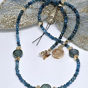 May include: A necklace with blue gemstone beads and gold-coloured details. The necklace has a decorative pendant and clover-shaped clasps. The beads are a deep blue colour, and the necklace is displayed on a white surface.