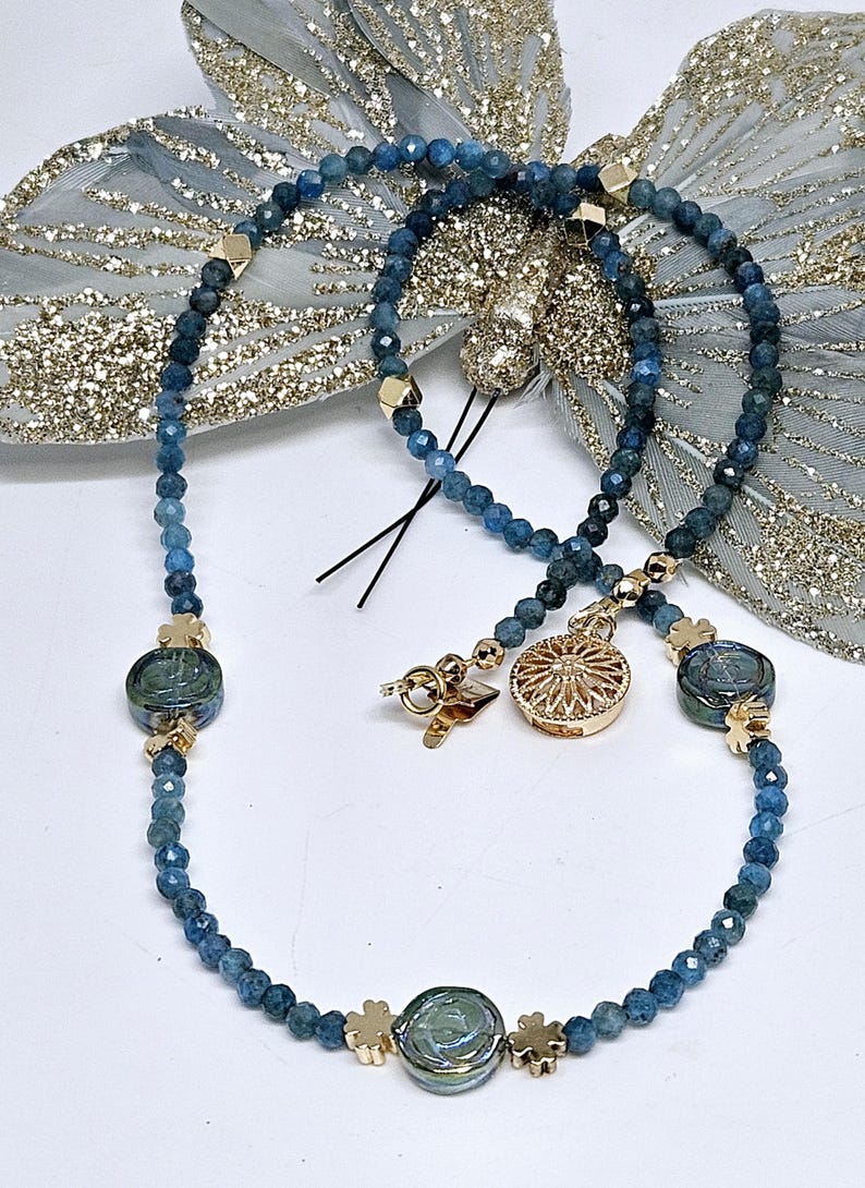May include: A necklace with blue gemstone beads and gold-coloured details. The necklace has a decorative pendant and clover-shaped clasps. The beads are a deep blue colour, and the necklace is displayed on a white surface.