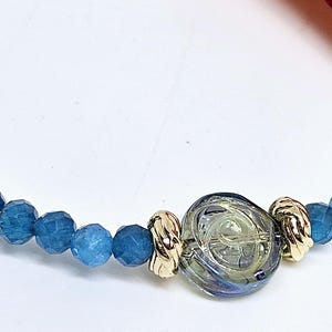 May include: A bracelet with faceted blue beads and gold-coloured metal accents. It features a central, clear glass bead with a swirling design. The bracelet is approximately 18 cm in length.