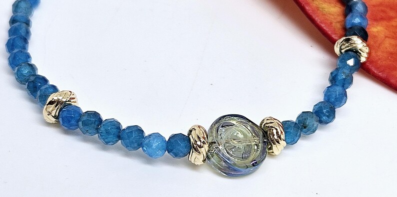 May include: A bracelet with faceted blue beads and gold-coloured metal accents. It features a central, clear glass bead with a swirling design. The bracelet is approximately 18 cm in length.