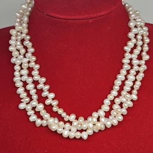 May include: A multi-strand pearl necklace displayed on a red velvet bust. The necklace features several strands of off-white, irregularly shaped pearls. The pearls are strung together, creating a layered effect. The background is a solid red.