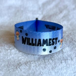 May include: A light blue fabric collar featuring the black text WILLIAMEST, small black paw prints, and cartoon dog illustrations. The collar is formed into a circular loop, showcasing the repeating pattern printed on the smooth material.