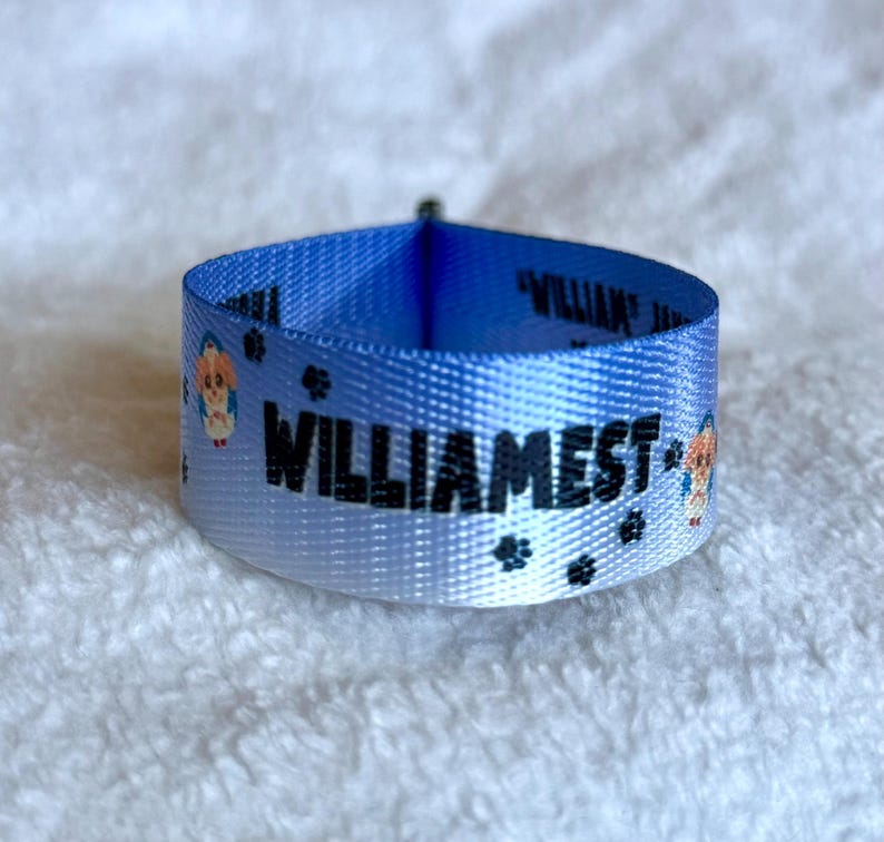 May include: A light blue fabric collar featuring the black text WILLIAMEST, small black paw prints, and cartoon dog illustrations. The collar is formed into a circular loop, showcasing the repeating pattern printed on the smooth material.