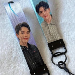 May include: Two fabric wristlet lanyards featuring printed portraits of individuals. One lanyard has a purple background with a person in a black shirt, while the other has a light blue background with a person in a grey shirt and a small colorful graphic.