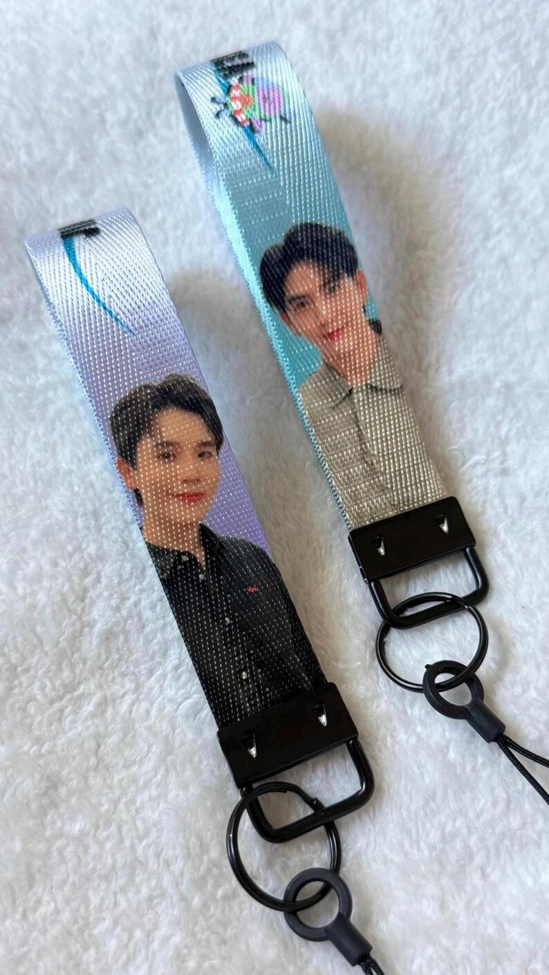 May include: Two fabric wristlet lanyards featuring printed portraits of individuals. One lanyard has a purple background with a person in a black shirt, while the other has a light blue background with a person in a grey shirt and a small colorful graphic.
