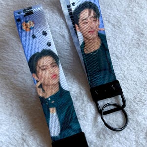 May include: Two fabric wristlet keychains featuring a blue gradient background with small black paw prints and a circular icon. Each strap displays a photographic portrait of a person with dark hair wearing a dark green shirt, attached to a black metal clasp and ring.