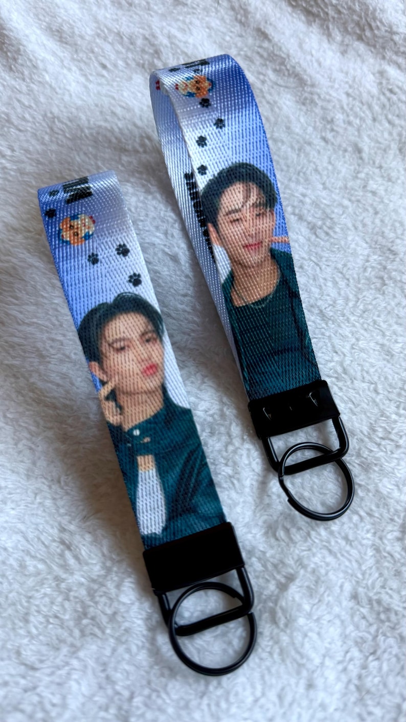 May include: Two fabric wristlet keychains featuring a blue gradient background with small black paw prints and a circular icon. Each strap displays a photographic portrait of a person with dark hair wearing a dark green shirt, attached to a black metal clasp and ring.
