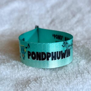 May include: A teal fabric wristband featuring the black text PONDPHUWIN printed in a bold font. The wristband includes a small blue cartoon character graphic, musical note symbols, and additional text that reads POND PHUWIN in black ink.