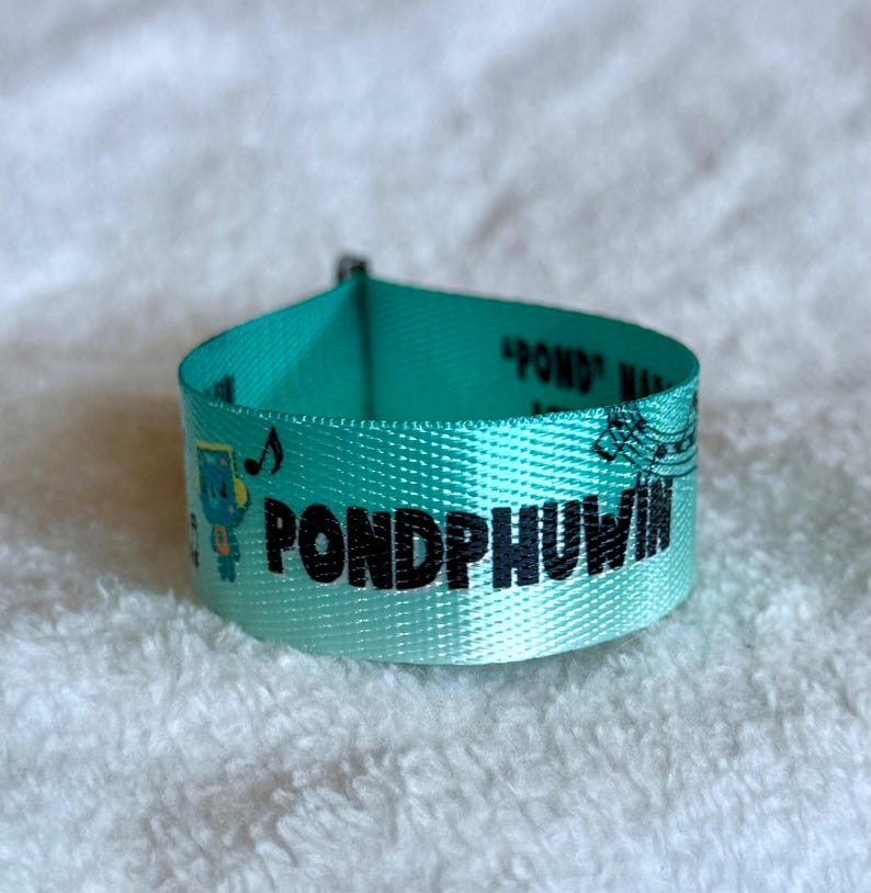 May include: A teal fabric wristband featuring the black text PONDPHUWIN printed in a bold font. The wristband includes a small blue cartoon character graphic, musical note symbols, and additional text that reads POND PHUWIN in black ink.