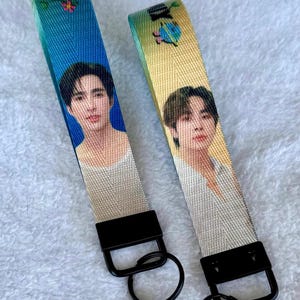May include: Two fabric wristlet keychains with black metal hardware and circular rings. One strap features a blue gradient background with a portrait of a person, while the other has a yellow gradient background with a different person's portrait.