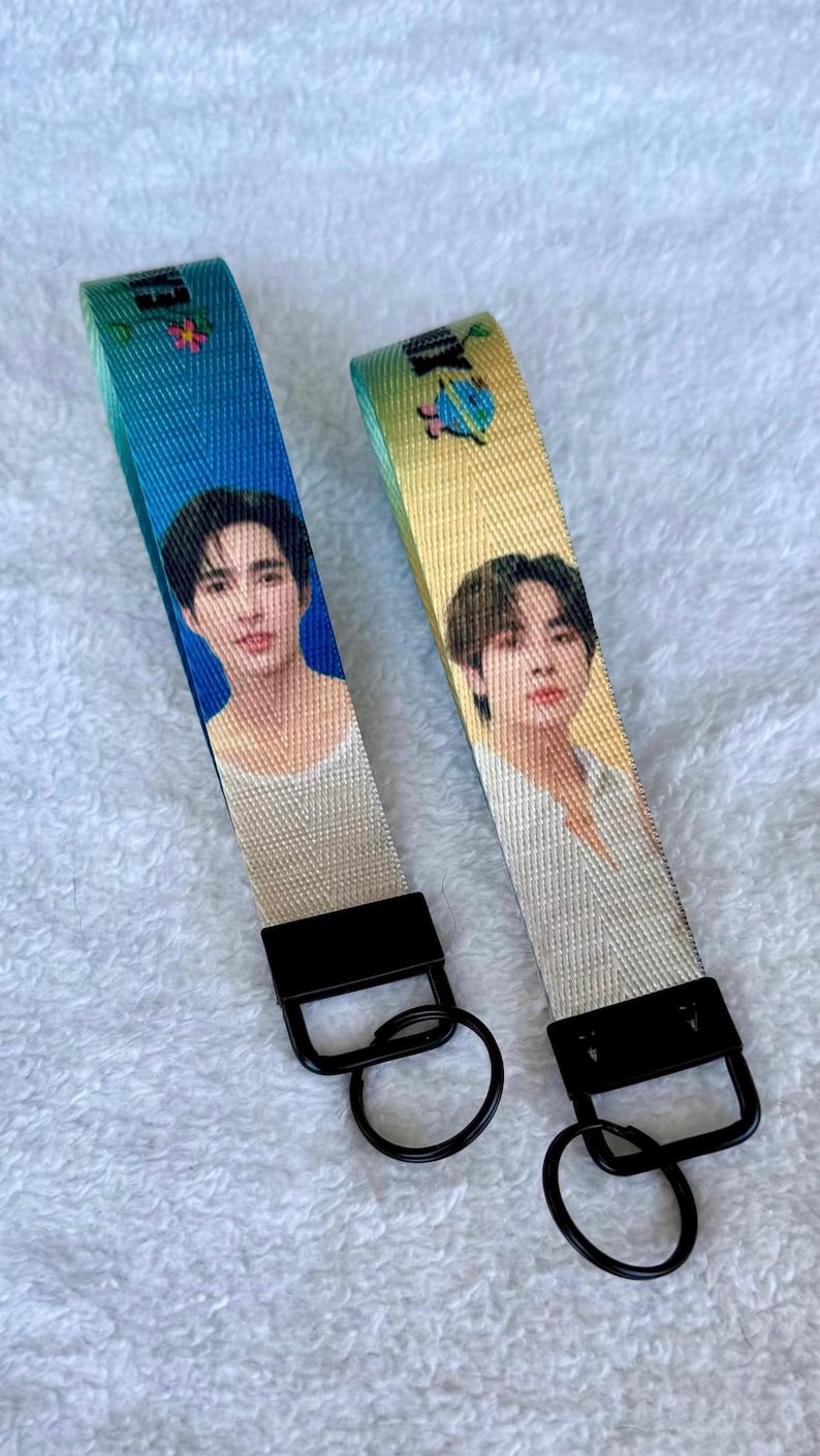 May include: Two fabric wristlet keychains with black metal hardware and circular rings. One strap features a blue gradient background with a portrait of a person, while the other has a yellow gradient background with a different person's portrait.