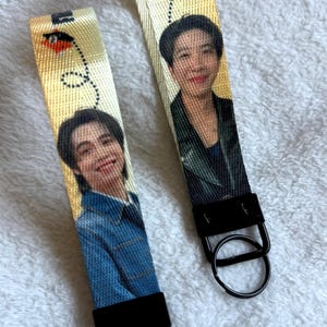 May include: Two fabric wristlet keychains featuring a yellow background with a repeating kite graphic and a photographic portrait of a person. Each keychain has a black metal clasp and a circular black metal split ring for keys.