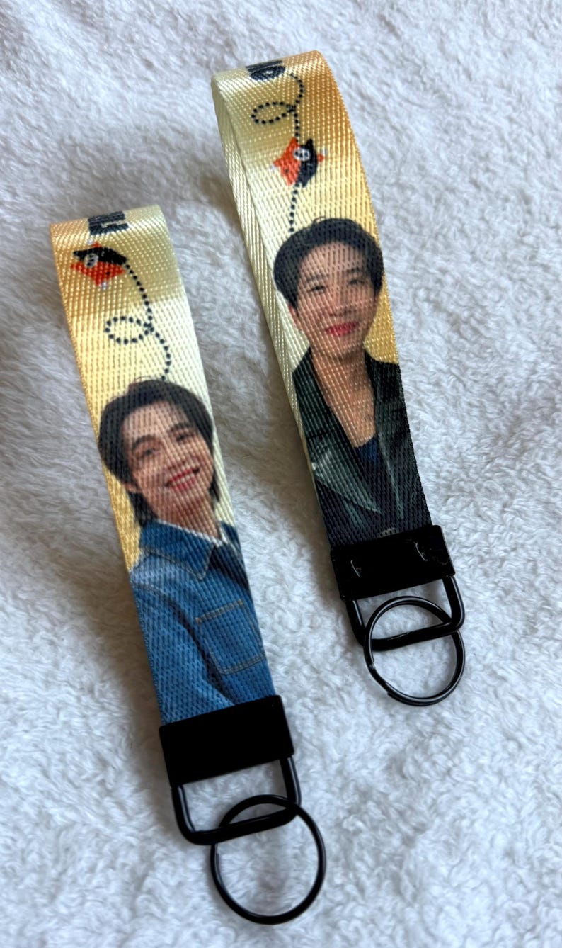 May include: Two fabric wristlet keychains featuring a yellow background with a repeating kite graphic and a photographic portrait of a person. Each keychain has a black metal clasp and a circular black metal split ring for keys.