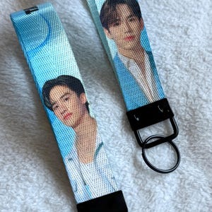 May include: Two blue fabric wristlet keychains featuring a printed portrait of a person with dark hair wearing a white shirt. Each strap is attached to a black metal clasp and a circular black metal key ring, creating a functional accessory.