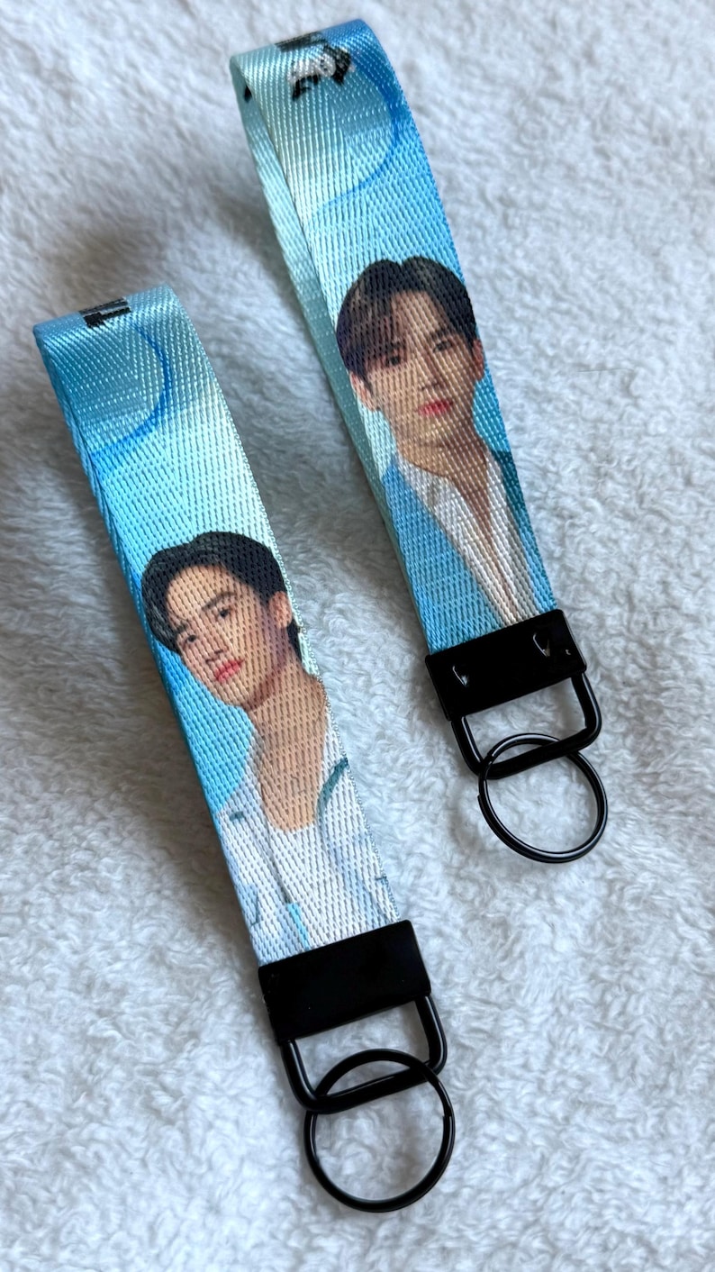 May include: Two blue fabric wristlet keychains featuring a printed portrait of a person with dark hair wearing a white shirt. Each strap is attached to a black metal clasp and a circular black metal key ring, creating a functional accessory.