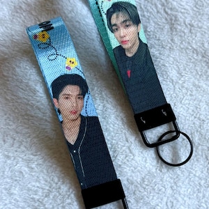 May include: Two fabric wristlet keychains featuring a printed portrait of a person with dark hair wearing a black shirt. The light blue straps are decorated with small yellow flower icons and black dashed lines, finished with black metal hardware and rings.