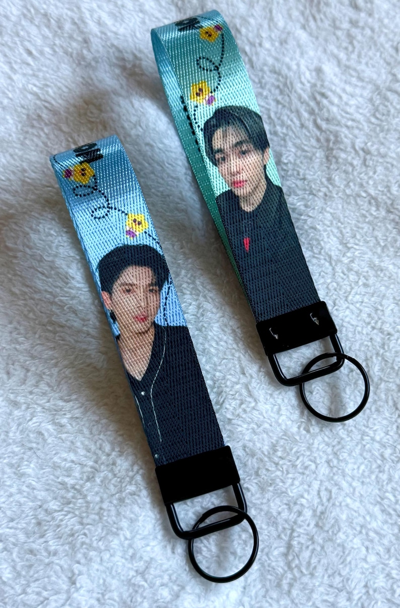 May include: Two fabric wristlet keychains featuring a printed portrait of a person with dark hair wearing a black shirt. The light blue straps are decorated with small yellow flower icons and black dashed lines, finished with black metal hardware and rings.