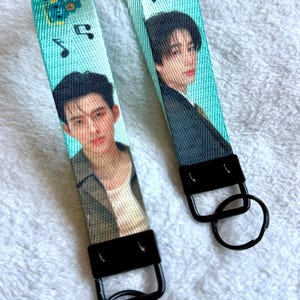 May include: Two fabric wristlet keychains in a bright turquoise color, each featuring a printed portrait of a person with dark hair. The straps are decorated with black musical note graphics and are attached to black metal hardware with round key rings.