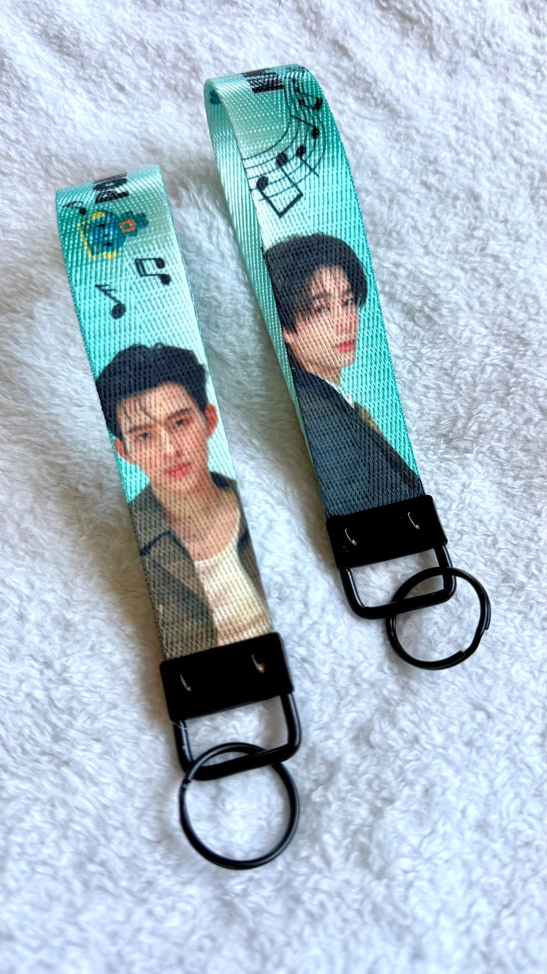 May include: Two fabric wristlet keychains in a bright turquoise color, each featuring a printed portrait of a person with dark hair. The straps are decorated with black musical note graphics and are attached to black metal hardware with round key rings.