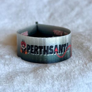 May include: A woven fabric label with a gray to black gradient background. The text PERTHSANTA is embroidered in black, with a small red devil tail extending from the letter A. A small graphic of a red devil character is positioned to the left.