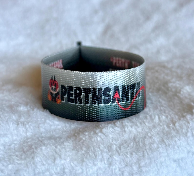 May include: A woven fabric label with a gray to black gradient background. The text PERTHSANTA is embroidered in black, with a small red devil tail extending from the letter A. A small graphic of a red devil character is positioned to the left.