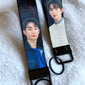 May include: Two black fabric wristlet keychains featuring printed portraits of individuals. Each strap has a black metal clasp and a circular black metal ring attached at the bottom. The fabric displays a repeating pattern of red dots.