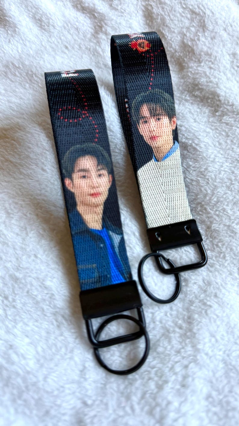 May include: Two black fabric wristlet keychains featuring printed portraits of individuals. Each strap has a black metal clasp and a circular black metal ring attached at the bottom. The fabric displays a repeating pattern of red dots.