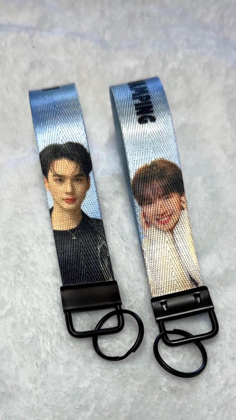 May include: Two light blue fabric keychains featuring printed portraits of individuals. Each strap has a black metal clasp and a circular black keyring. The right strap displays the text JUMPING in black letters at the top, while the left strap shows a person in a black shirt.