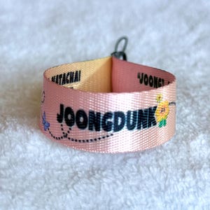 May include: A pink fabric wristband featuring the black text JOONGDUNK, the name NETACHAI, and a small yellow flower graphic with a blue butterfly. The band is looped and secured with a small black metal ring at the top, displaying a smooth texture.