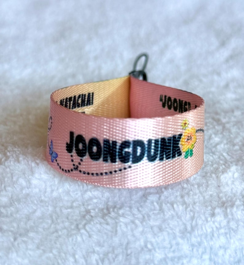 May include: A pink fabric wristband featuring the black text JOONGDUNK, the name NETACHAI, and a small yellow flower graphic with a blue butterfly. The band is looped and secured with a small black metal ring at the top, displaying a smooth texture.