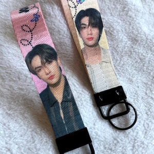 May include: Two fabric wristlet keychains featuring printed portraits of a person with dark hair. The left strap is pink with a black jacket portrait, and the right is cream with a light-colored shirt portrait. Both have black hardware and rings.
