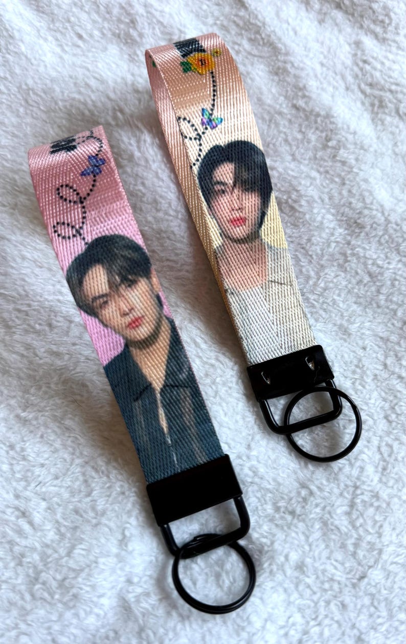 May include: Two fabric wristlet keychains featuring printed portraits of a person with dark hair. The left strap is pink with a black jacket portrait, and the right is cream with a light-colored shirt portrait. Both have black hardware and rings.