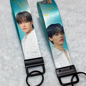 May include: Two fabric wristlet keychains featuring a teal background with yellow star accents and a photographic print of a person with dark hair wearing a white shirt. Each keychain has a black metal clasp and a circular black metal key ring.