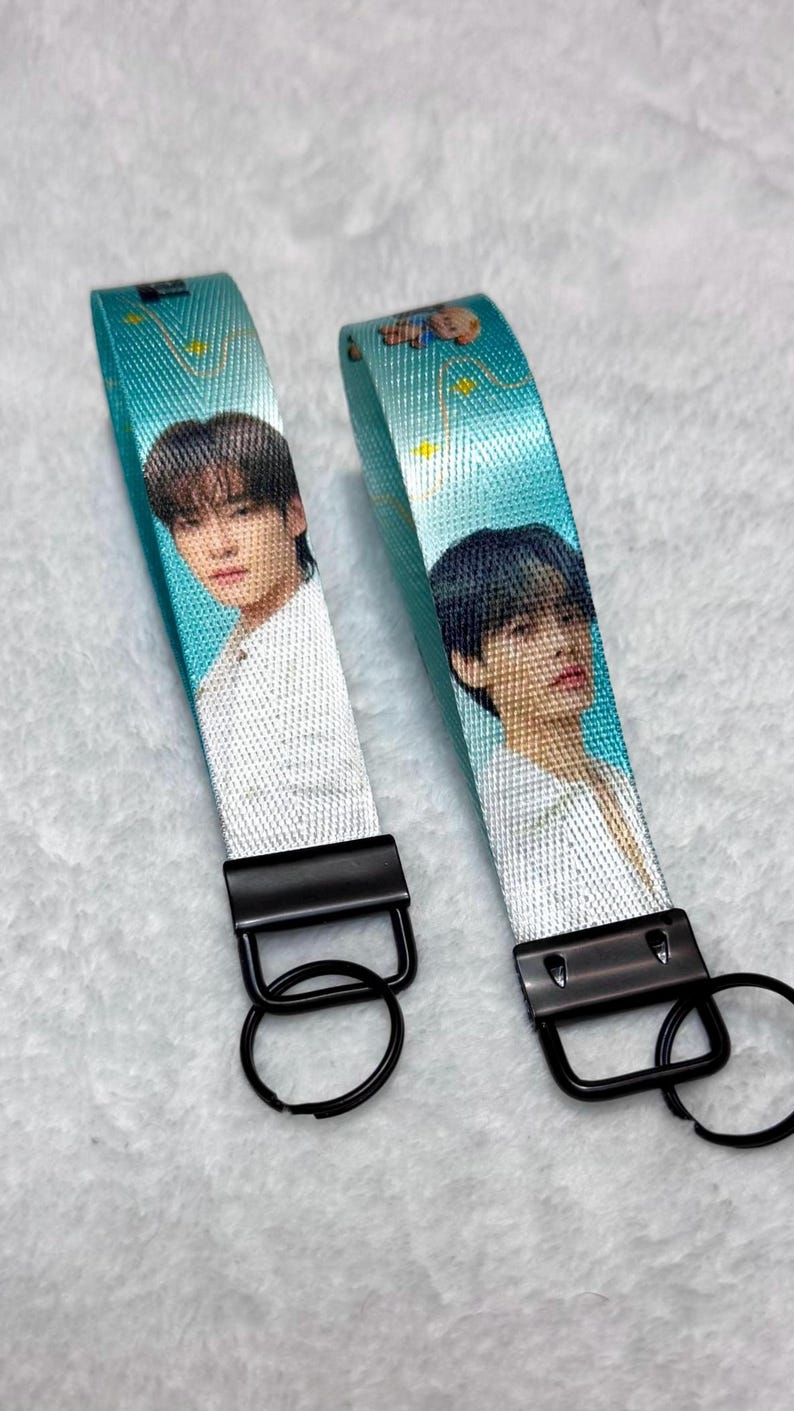 May include: Two fabric wristlet keychains featuring a teal background with yellow star accents and a photographic print of a person with dark hair wearing a white shirt. Each keychain has a black metal clasp and a circular black metal key ring.