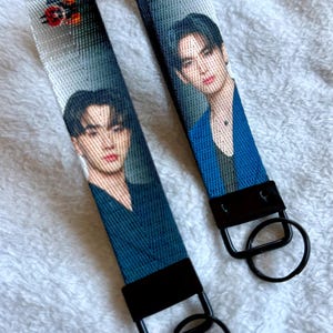 May include: Two fabric wristlet keychains featuring a printed portrait of a person with dark hair. Each strap is made of a woven material with a black metal clasp and a circular black split ring attached at the bottom for holding keys.
