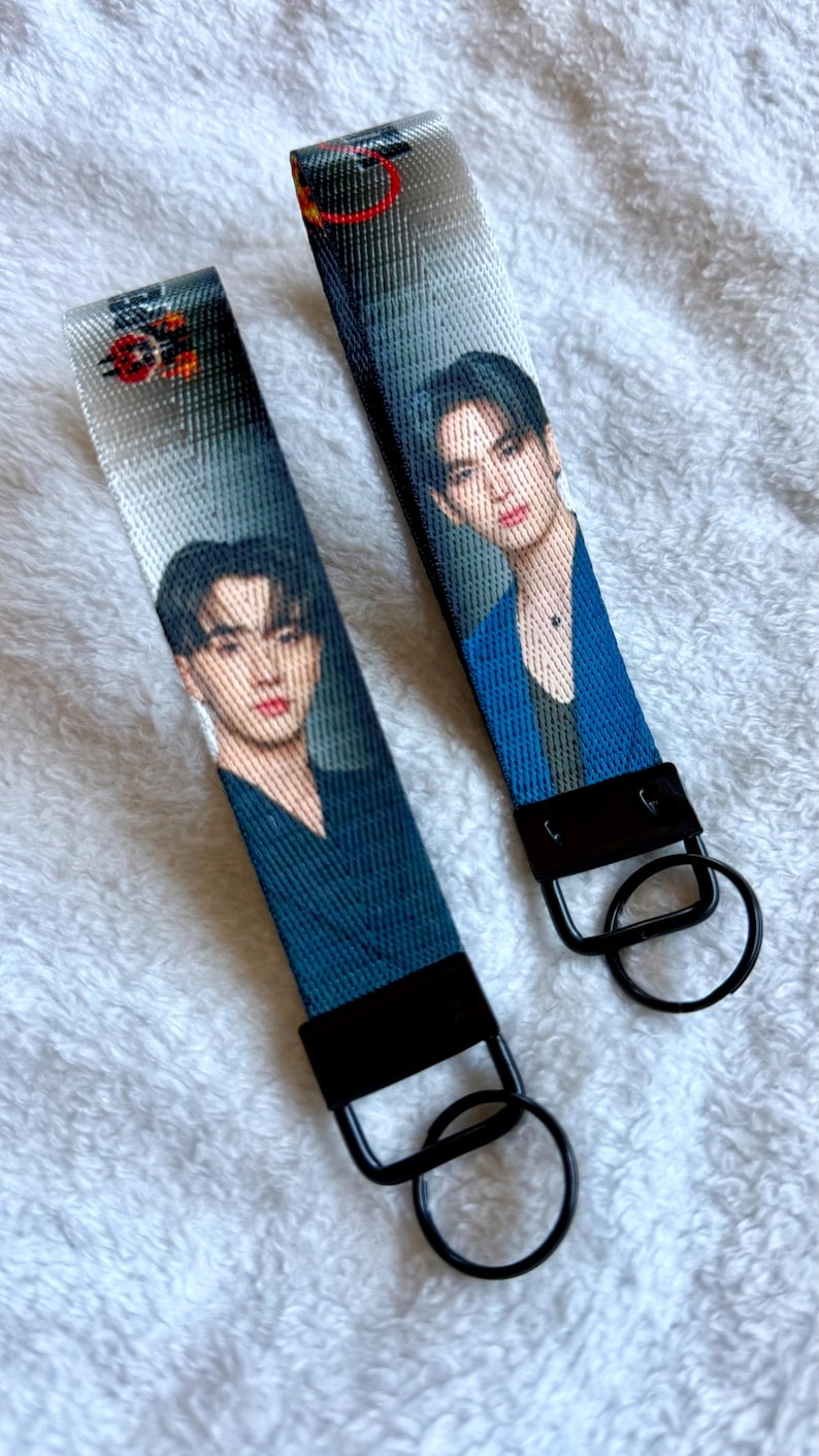 May include: Two fabric wristlet keychains featuring a printed portrait of a person with dark hair. Each strap is made of a woven material with a black metal clasp and a circular black split ring attached at the bottom for holding keys.