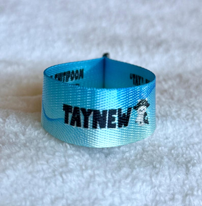 May include: A light blue fabric wristband featuring a gradient color effect. The text TAYNEW is printed in bold black letters on the front, accompanied by a small graphic of a character wearing a hat. The band has a smooth, woven texture.