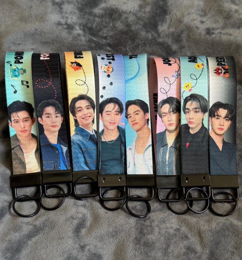 May include: A set of eight fabric wristlet keychains featuring printed portraits of individuals in various colored backgrounds. Each strap includes a black metal clasp and a circular key ring. The designs incorporate small graphic icons and text.