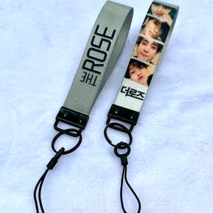 May include: Two wrist straps. One is gray with "THE ROSE" in black. The other is white with images of people and Korean text. Both have black hardware and black cords.