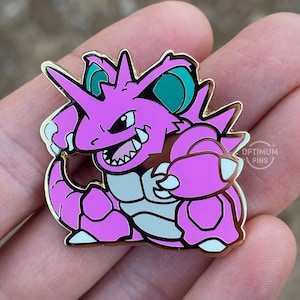May include: A pink and teal enamel pin depicting a horned creature with sharp teeth, set within a gold border. The pin includes the text "OPTIMUM PINS". The creature is in a dynamic pose. The design is vibrant and detailed.