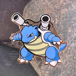 May include: A blue and tan pin featuring a cartoon Blastoise character with two cannons on its back. The pin has a gold outline and the text "OPTIMUM PINS" is visible.