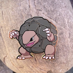 May include: A pin featuring a cartoon character with a gray, rock-like body and a tan face and limbs. The character has a determined expression and is outlined in gold. The pin is set against a large, gray stone.