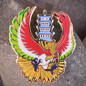 May include: A gold-colored enamel pin featuring a red and white bird with green and yellow accents. The bird is flying with its wings spread and has a pagoda behind it. The pin has the text "OPTIMUM PINS" on the bottom.