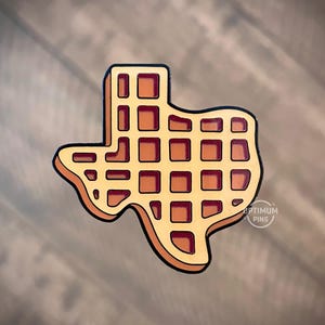 May include: A pin in the shape of the state of Texas, designed to look like a waffle. The pin is light brown with a darker brown outline and waffle grid pattern. The text "OPTIMUM PINS" is visible on the bottom right.
