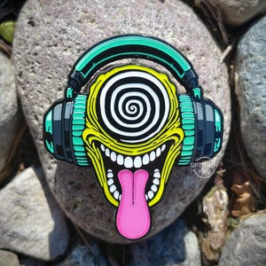 Pode incluir: Um pin colorido de esmalte com um monstro de desenho animado com fones de ouvido. O monstro tem um rosto amarelo, uma língua rosa e um olho espiral em preto e branco. Os fones de ouvido são verde-azulado e preto.