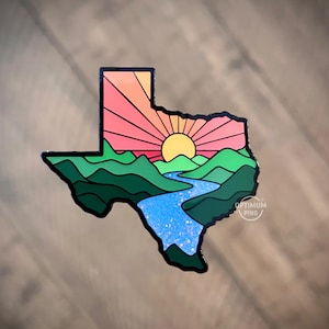 Könnte beinhalten: Texas-förmiger Ansteckpin mit Sonnenuntergangsdesign, gelber Sonne und strahlenden Orange- und Rosatönen. Der Pin zeigt grüne Hügel, einen blauen Fluss und den Text "OPTIMUM PINS".