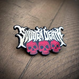 Könnte beinhalten: Ein schwarz-weißer Emaille-Pin mit dem Schriftzug "Sudden Death" über drei roten Totenköpfen. Der Pin hat einen schwarzen Rand und ist vor einem Holzgrund platziert. Der Pin ist etwa 3,8 cm breit.
