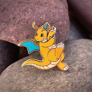 May include: A gold-coloured Dragonite enamel pin with a light orange body, blue wings, and white accents. The pin features the Dragonite character in a playful pose. The pin is set against a background of dark brown and grey rocks. The text "OPTIMUM PINS" is visible.